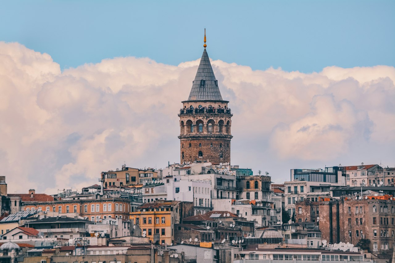 The stone Galata Tower and its conical roof rising above the crowded rooftops of Istanbul's Galata quarter against a bank of white cloud